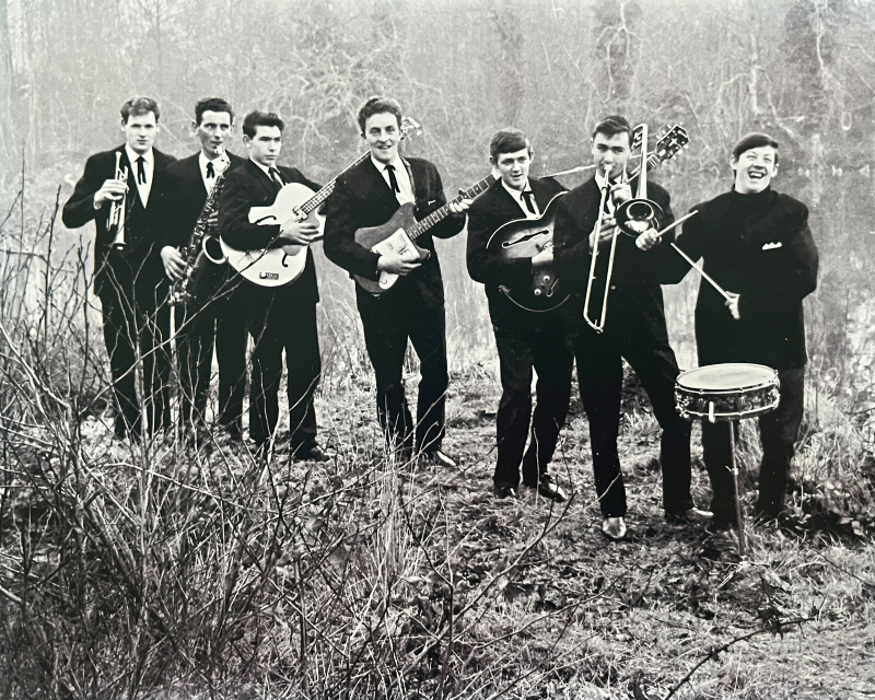 Black and white photo of the Embassy Showband, circa 1960s. Tom Foley's dad playing guitar at center. The seven band members are dressed in suits and ties, several smiling widely, standing in a field clearing.