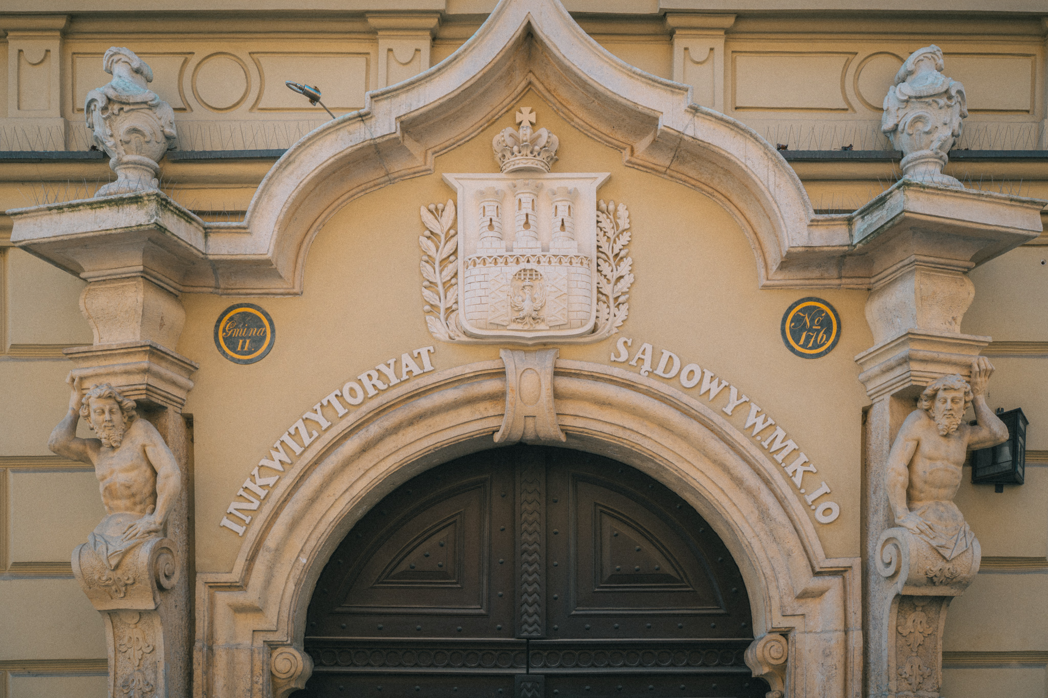 Facade from Kraków City Center. Above the wooden arched doorway, serif letters are carved in all caps in relief, with a crest and other decorative statues and carvings framing it.