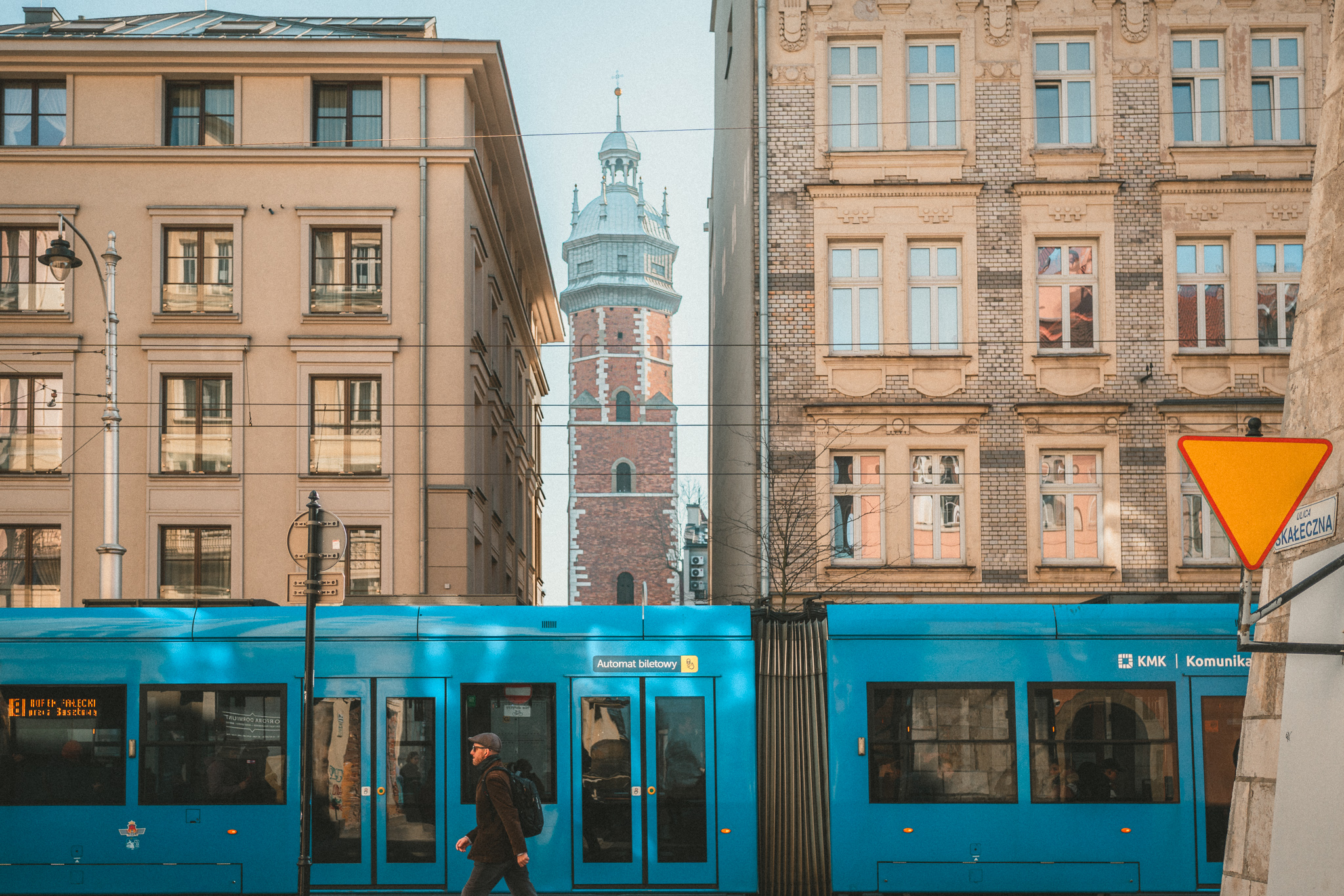 A street scene in Kazimierz in Kraków. A teal bus drives past historic buildings as a pedestrian dressed in dark colored clothing with a black backpack walks past.