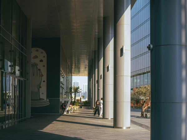 Image of two people walking under a large building's overhang, between columns to the sunny sidewalk. A mural of a upfaced hand with small clouds in different colors is painted on the building. People sit outside at tables, looking at the sunshine.