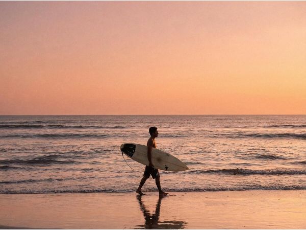 A surfer carrying a surfboard on the beach at sunset in Kuta.