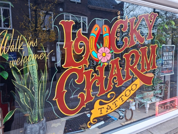 The window of a tattoo shop features nadpainted signage in English with its name, and declaring "walk-ins welcome!"