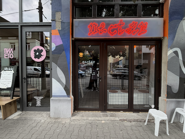 Pileto, at left, a chicken restaurant with logo of a pastel pink circle with a cartoon rooster head in the middle in black. Neon signage on right.