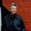 Spike Spondike portrait, wearing a black jacket over a black button-down with a black bandanna tied around her neck, in front of a rust-red wall.