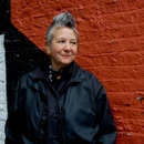 Spike Spondike portrait, wearing a black jacket over a black button-down with a black bandanna tied around her neck, in front of a rust-red wall. Spike Spondike portrait, wearing a black jacket over a black button-down with a black bandanna tied around her neck, in front of a rust-red wall.