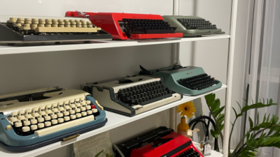 Typewriters on three shelves in a variety of colors (red, teal, grey with white or black keys).