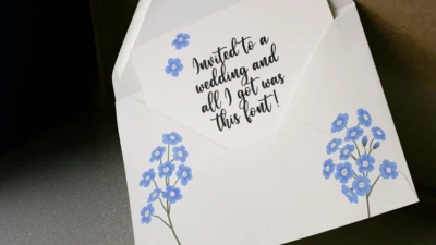 A white envelope with blue flowers containing a white notecard with two blue flowers and the text: "Invited to a wedding and all I got was this font!" The font used for the text is a stereotypical wedding font.