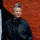 Spike Spondike portrait, wearing a black jacket over a black button-down with a black bandanna tied around her neck, in front of a rust-red wall. Spike Spondike portrait, wearing a black jacket over a black button-down with a black bandanna tied around her neck, in front of a rust-red wall.
