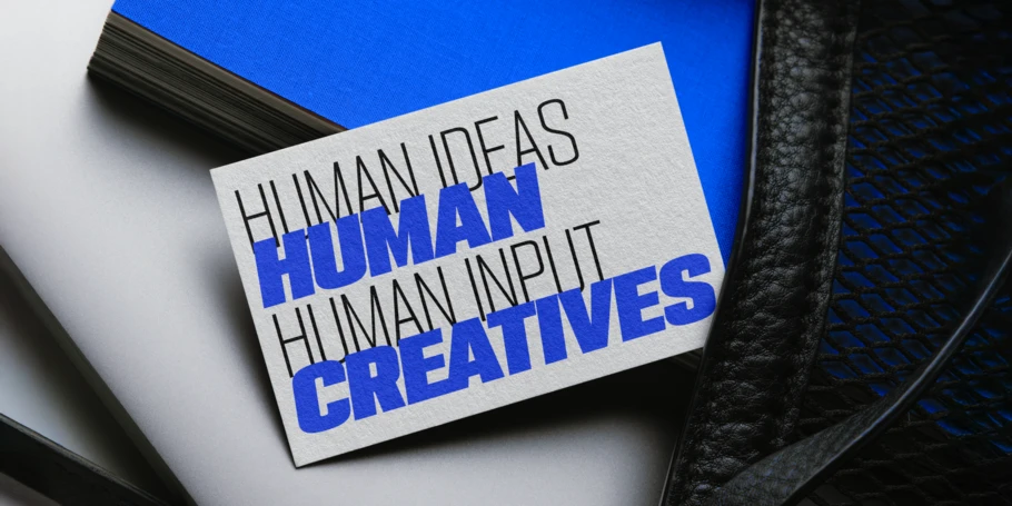 Bright blue notebook on top of a silver laptop, all in a bag's mesh pocket. A piece of paper with text printed is on top. Thin black text: "HUMAN IDEAS HUMAN INPUT" Thick blue text: "HUMAN CREATIVES"
